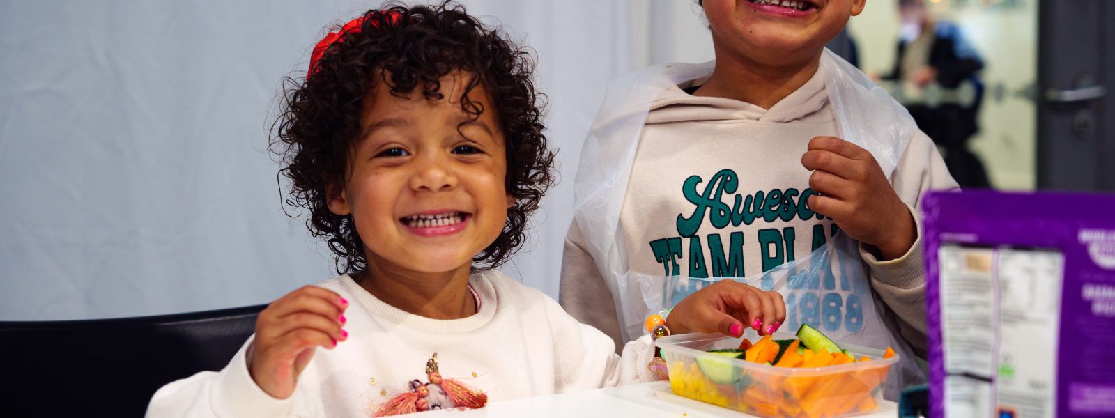 A little girl smiles at the camera as she eats fresh vegetables from a Tupperware.
